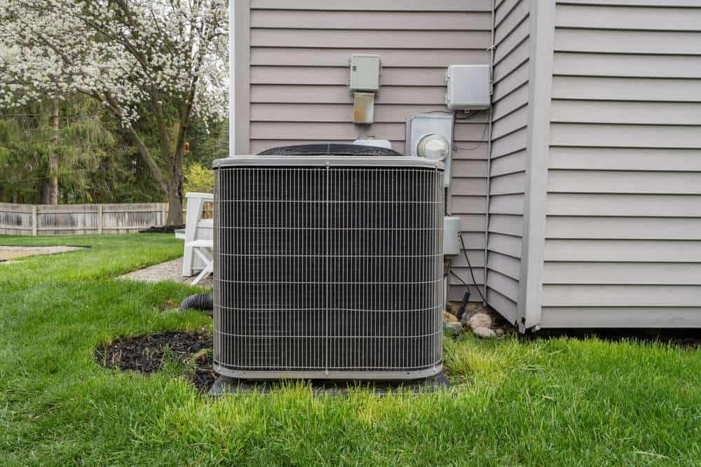 An outdoor air conditioning unit, recently installed as part of an AC installation project, is positioned on the lawn next to the exterior wall of a house. Nearby, a blooming tree and fence add to the serene setting.