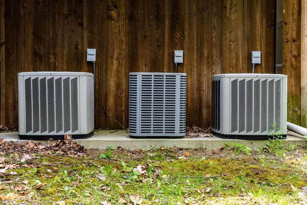 Three outdoor HVAC units are installed on a concrete pad against a wooden wall, with fallen leaves and grass on the ground around them. Regular AC maintenance, including air duct cleaning, ensures these units operate efficiently all year round.