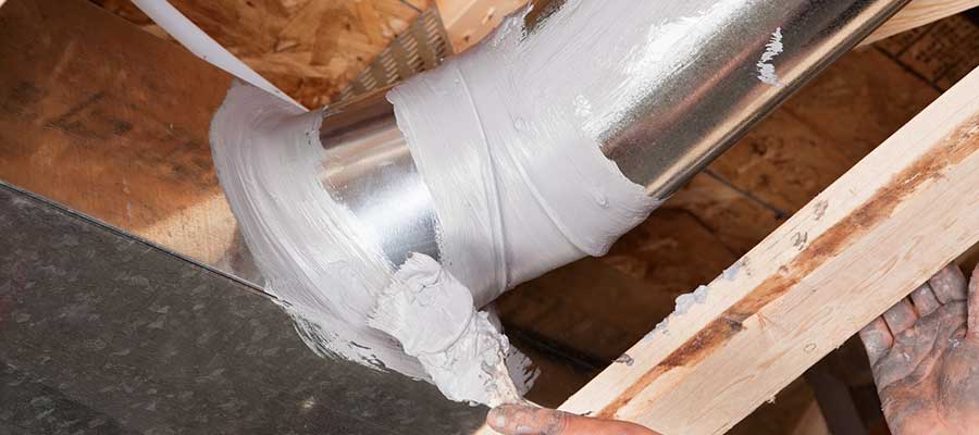 A person applies duct mastic sealant around an HVAC duct joint, ensuring an airtight seal within a wooden-framed ceiling space as part of a meticulous ac maintenance routine.