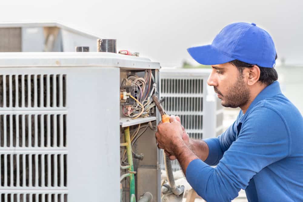 A technician in a blue cap and shirt is repairing an outdoor HVAC unit, focusing on the internal components with a tool in hand. His expertise extends beyond just repairs, including AC installation and air duct cleaning to ensure optimal performance.