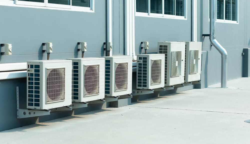 Several air conditioning units are mounted on an exterior wall of a building, beneath multiple windows. A drainpipe is visible on the right side, suggesting recent ac installation.