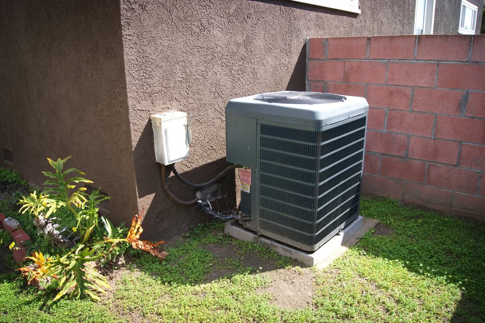 An outdoor air conditioning unit installed against a brown stucco wall, surrounded by a small grassy area and a few plants, hints of recent HVAC maintenance. A red brick wall is visible beside it.