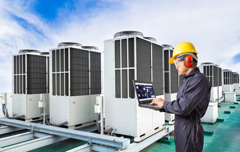 A technician in a yellow hard hat and protective gear uses a laptop to inspect and manage multiple rooftop HVAC units, ensuring efficient A/C repair under a clear blue sky.