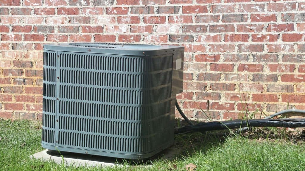 An outdoor air conditioning unit sits on a concrete slab next to a red brick wall, indicative of a recent ac installation. Black cables extend from the unit into the building, showcasing professional HVAC work.