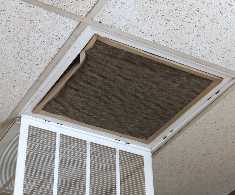 A ceiling air vent with the cover open reveals an old, dirty air filter inside, surrounded by speckled ceiling tiles. Proper HVAC maintenance, such as regular air duct cleaning and ac installation upgrades, can help prevent this buildup.