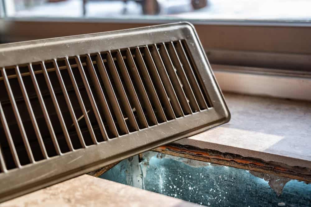 A floor vent grate has been removed, exposing the ductwork beneath it, likely during an a/c repair. The vent is placed at an angle on the floor near a window.