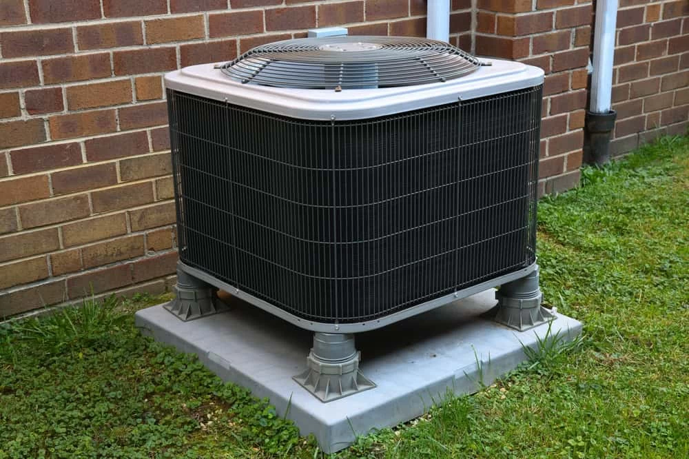 An outdoor air conditioning unit, ideal for those considering ac installation Sarasota, is positioned on a concrete pad next to a brick wall. Elevated on cylindrical footings and surrounded by grass and small plants, the metal casing and fan are visible on top.