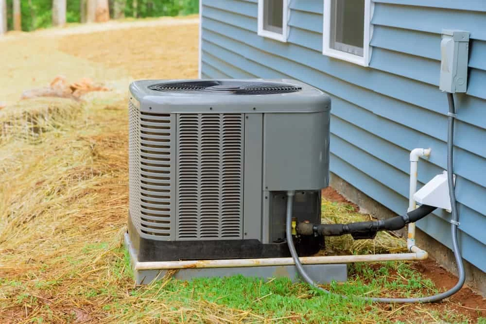 An outdoor air conditioning unit is positioned next to a blue house with vinyl siding, exemplifying expert air conditioning installation in Sarasota. The unit sits on a concrete slab, surrounded by dry grass and green patches, with electrical and refrigerant lines connecting it to the house.