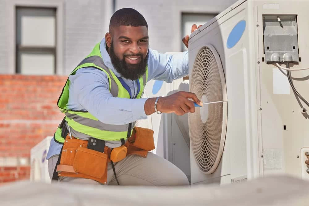 A smiling technician in a safety vest and tool belt expertly uses a screwdriver for air conditioning maintenance in Sarasota, ensuring the outdoor unit runs smoothly.